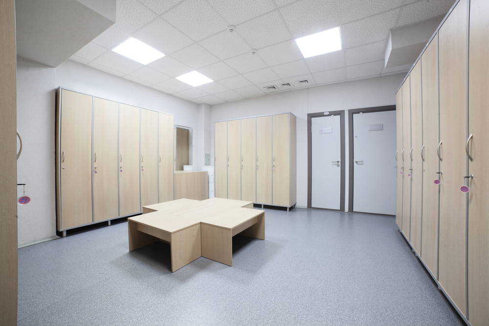 Locker room with tall wooden lockers along the walls, a central wooden seating table, and two gray doors at the back.