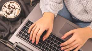 A person working on a laptop while sitting on the floor next to a hot drink.