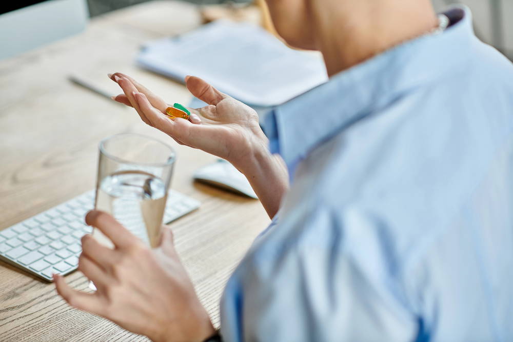 Person holds two colorful capsules in an open palm over a desk, with a glass of water, a keyboard, and papers nearby.