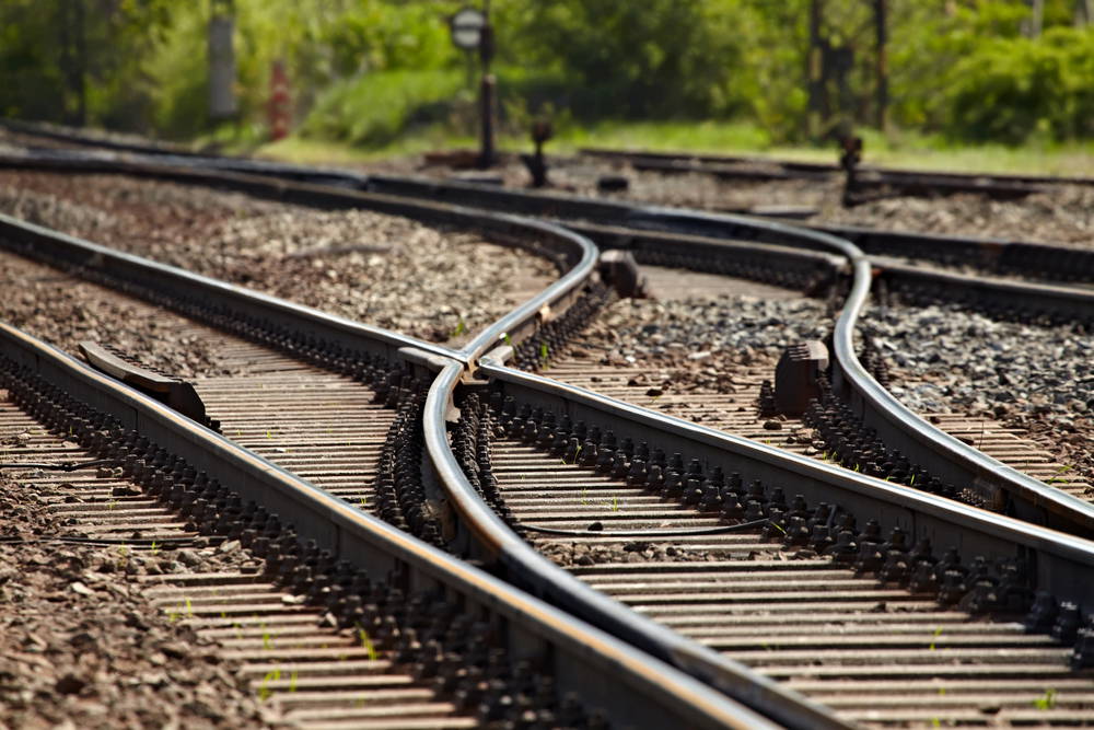 Railway tracks with multiple switch points diverging in a gravel yard, green trees in the blurred background.
