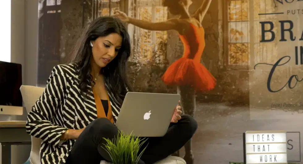 Woman in a striped blazer sits with a MacBook on her lap, in an office with a ballerina mural in the background.