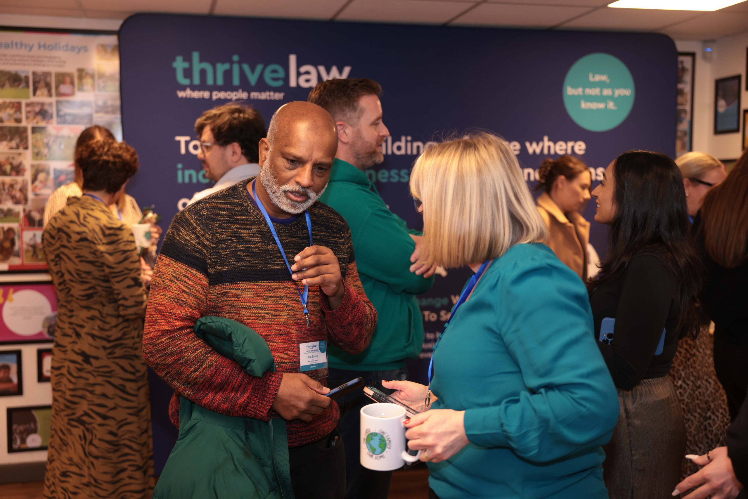 Bald man with a gray beard wearing a multicolor sweater talks with others at a Thrive Law event, holding a cup and a green jacket draped over his arm in front of a blue backdrop.