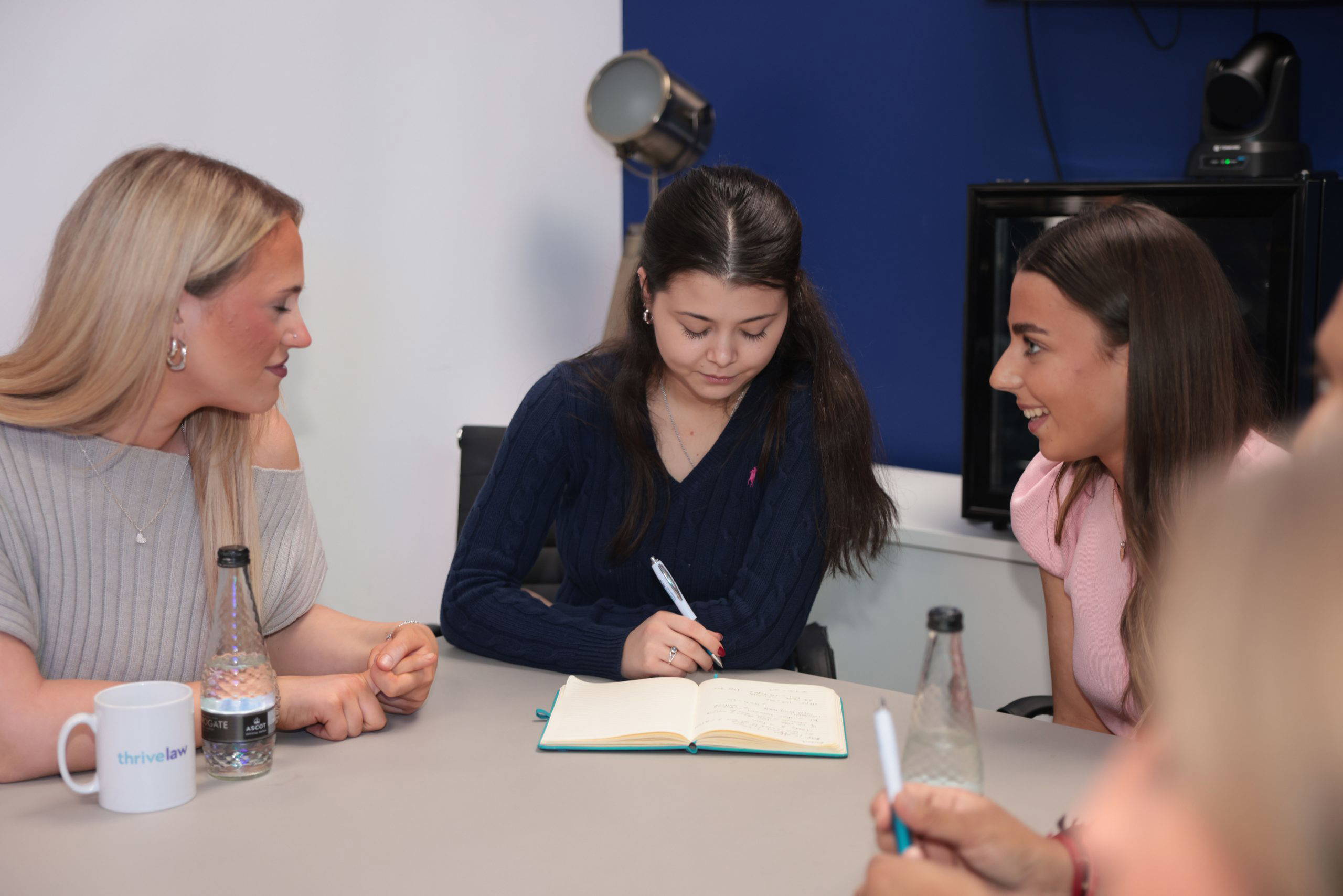 Three women sit around a table, writing in a notebook during a discussion in a bright office setting.