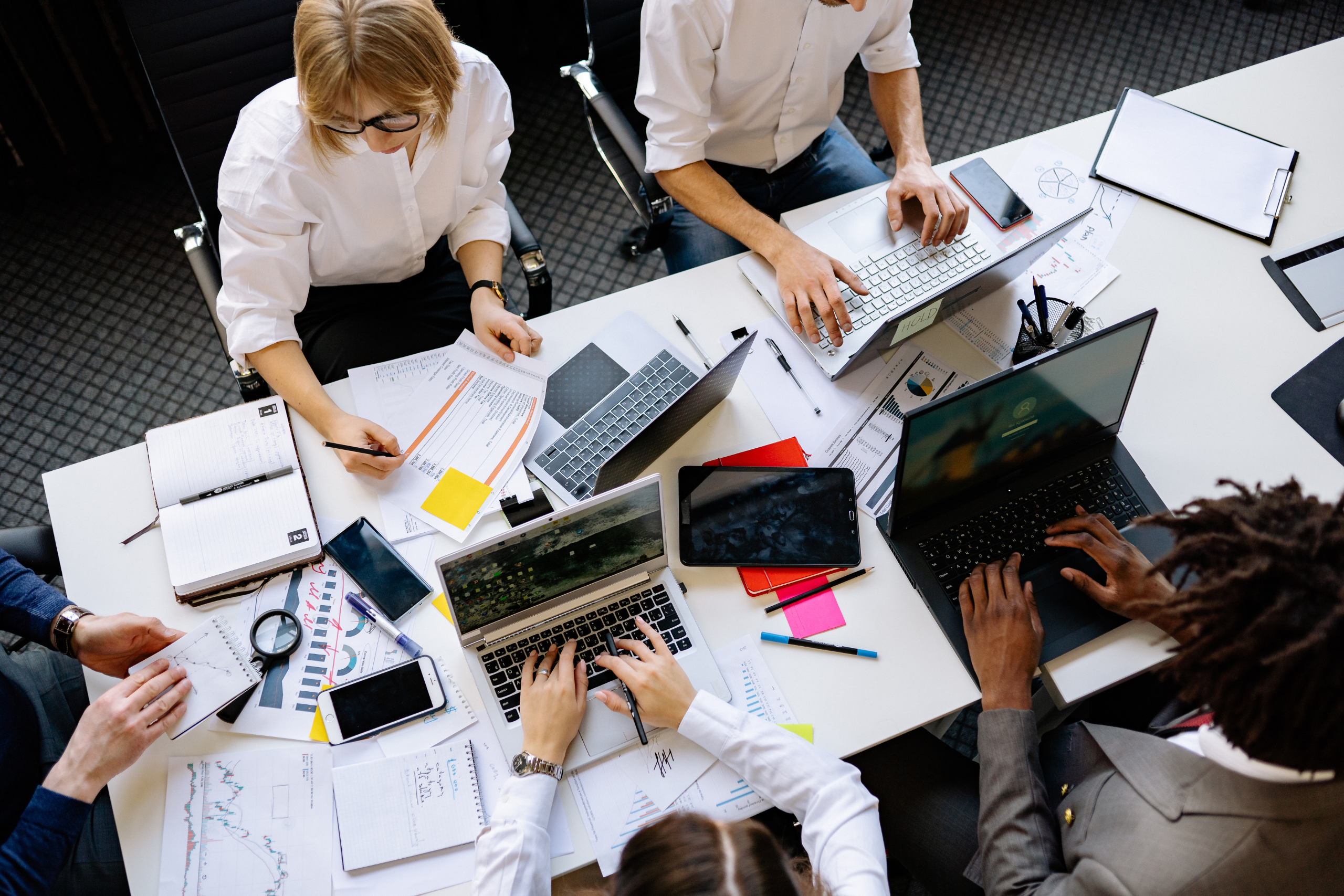 Four people working, sat at a desk featuring laptop computers and notebooks.