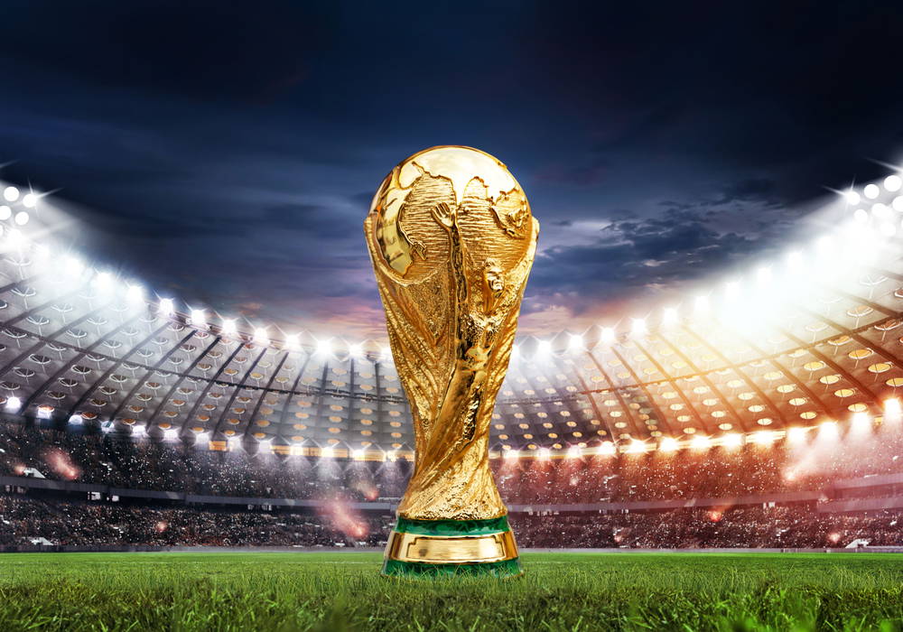Golden World Cup trophy standing on a grassy soccer field with a brightly lit stadium full of spectators in the background during dusk/night.