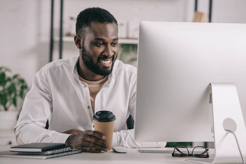 Man in a white shirt sits at a desk, smiling at a large desktop monitor with a coffee cup in hand.