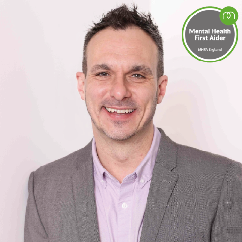 Smiling man in a gray blazer and lavender shirt, with a Mental Health First Aider badge in the top-right corner (MHFA England).