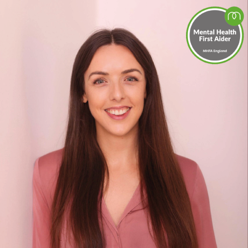 Portrait of a smiling woman with long brown hair wearing a pink blouse; Mental Health First Aider badge in the top-right corner.