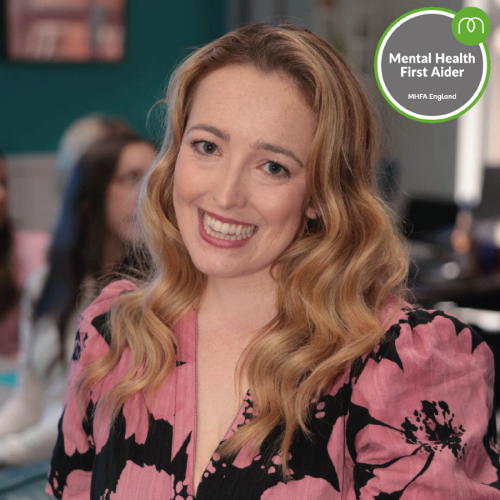 Smiling woman in a pink floral blouse in a workplace, with a Mental Health First Aider badge in the corner.