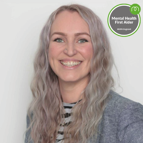 Smiling woman with wavy lavender hair wearing a striped top and gray blazer, MHFA England Mental Health First Aider badge in the corner